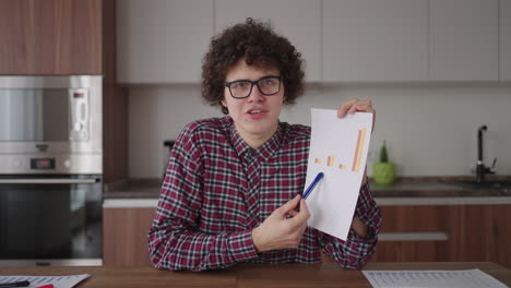 Curly---haired-with-glasses-business-man-sitting-at-office-from-home-desk-looking-at-camera-and-pointing-at-a-tablet-with-financial-information-displayed-in-graphical-form-column-graph